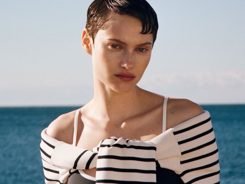 Close-up portrait of a short-haired woman wearing a striped sweater over her shoulders against an ocean horizon.