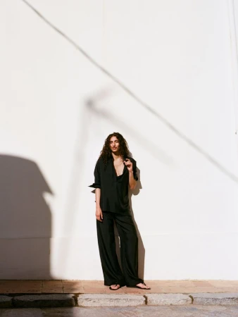 Model with long dark curly hair wearing black shirt and trousers leaning against a white building wall outdoors.