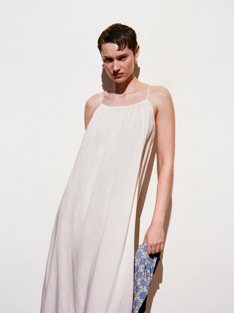 Model with short dark hair wearing a white slip dress holding a blue patterned fabric against a plain white wall