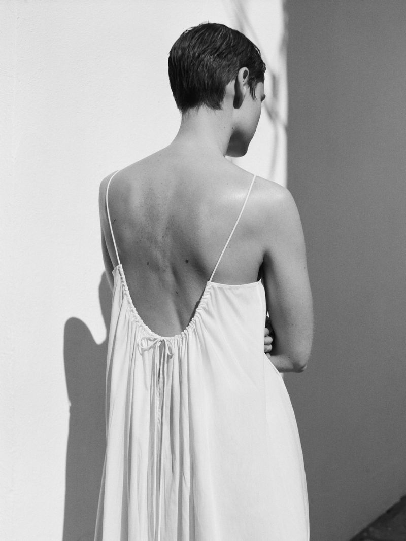 Model with short dark hair seen from behind wearing a white slip dress against a plain wall.