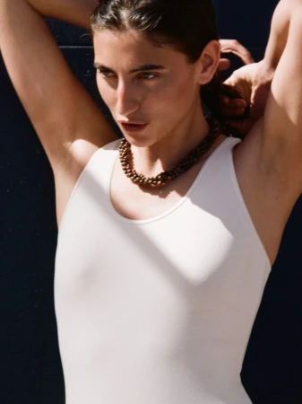Model wearing a white tank top and beaded necklace posing outdoors against a dark wall for Arket