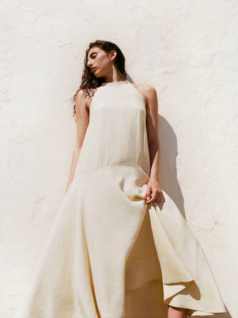 Model in a cream halterneck midi dress leaning against a textured off-white wall in bright natural sunlight.