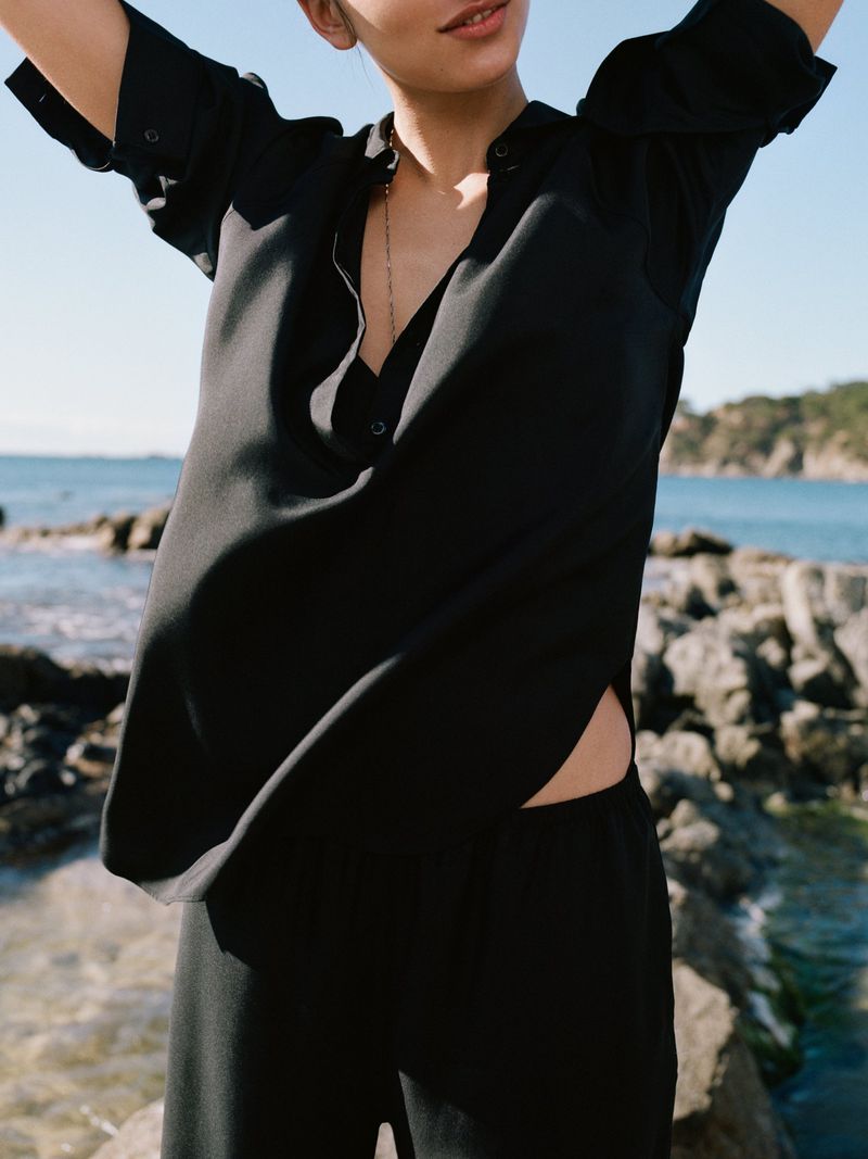 Model wearing a loose black button-down shirt outdoors near a rocky coastline and the ocean for the Arket S/S26 campaign.