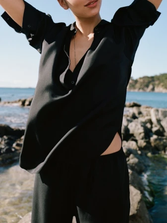 Model wearing a loose black button-down shirt outdoors near a rocky coastline and the ocean for the Arket S/S26 campaign.
