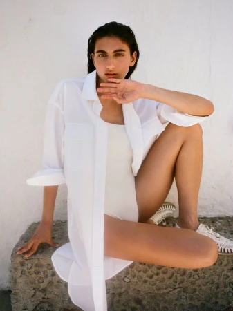 Woman in a white shirt and bodysuit seated on a low stone ledge against a white wall for the Arket S/S26 campaign.