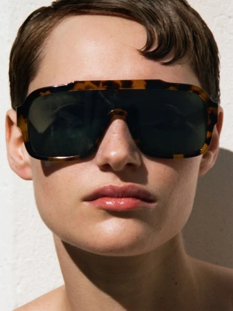 Close-up portrait of a model wearing tortoiseshell sunglasses against a white wall.