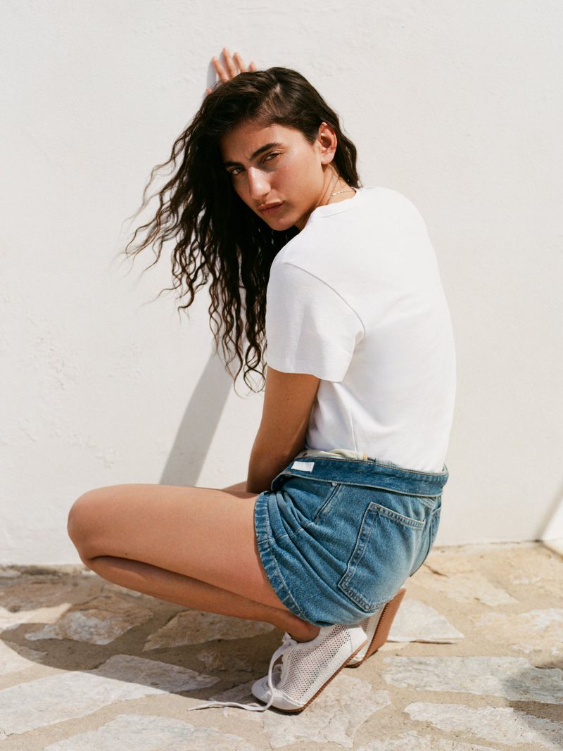 Model crouching in a white t-shirt and denim shorts against a light wall for Arket campaign