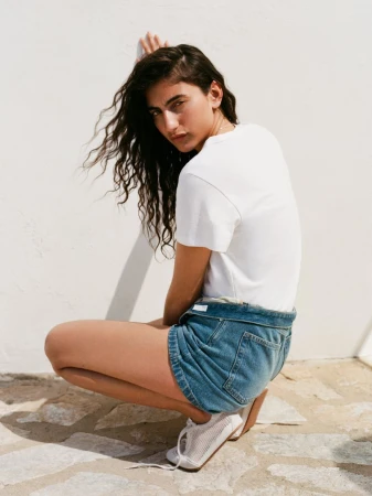 Model crouching in a white t-shirt and denim shorts against a light wall for Arket campaign