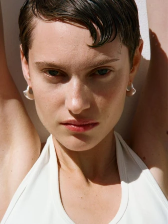 Close-up portrait of a woman with short dark hair wearing a white top and silver hoop earrings against a white background.