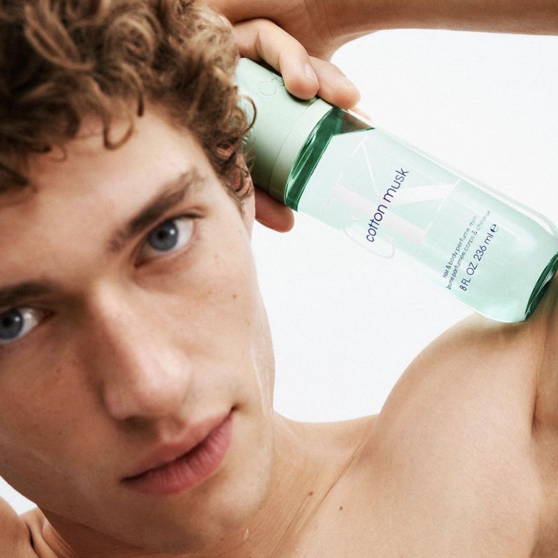 Close-up shot of a shirtless adult male model holding a green and white Cotton Musk spray bottle up to his head in a bright studio.