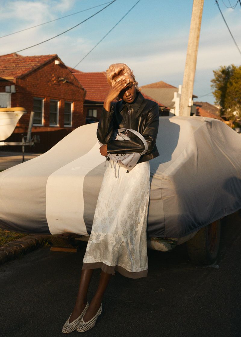 Adult female in a black jacket and floral skirt poses outside near a car covered with a grey tarp in natural light against a house and sky.