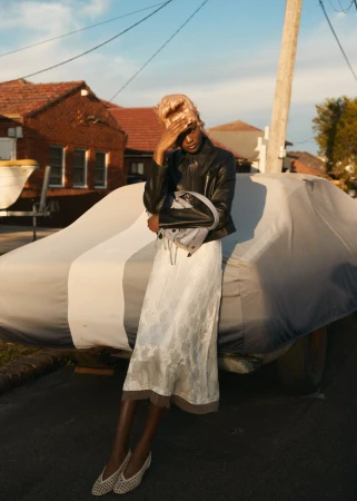 Adult female in a black jacket and floral skirt poses outside near a car covered with a grey tarp in natural light against a house and sky.