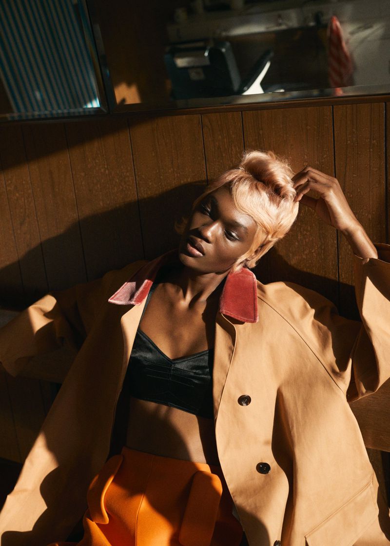 Model Naki Depass in a camel coat, black crop top, and orange trousers posed against a wood-paneled wall in dramatic sunlight.