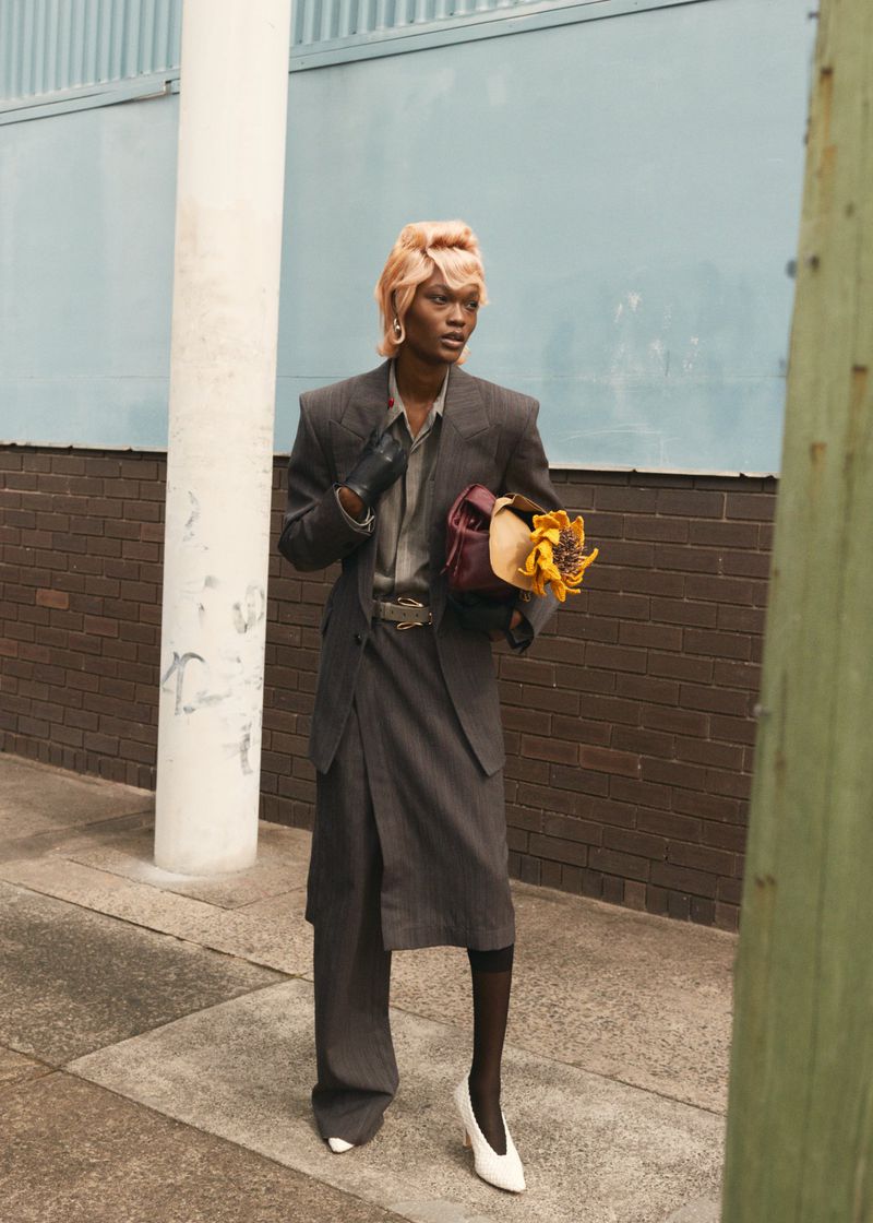 Elegant non-binary model poses outdoors in oversized grey suit, carrying flowers and handbag, wearing white heels, against brick wall.