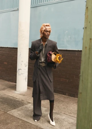Elegant non-binary model poses outdoors in oversized grey suit, carrying flowers and handbag, wearing white heels, against brick wall.