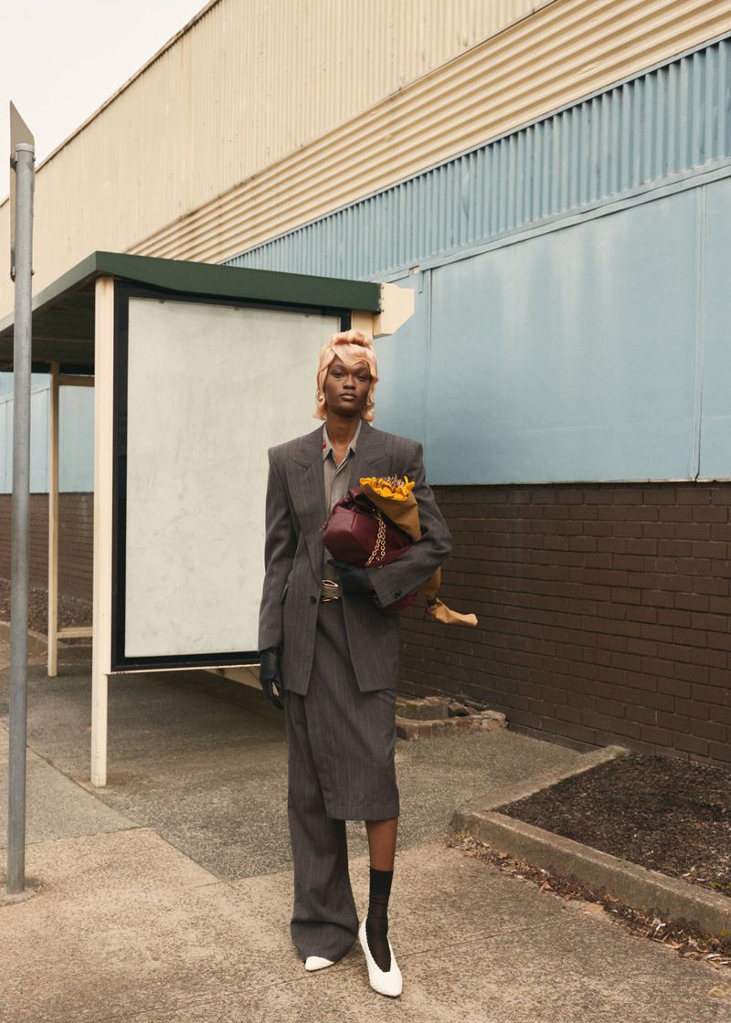 Woman in grey suit stands by bus stop holding bag and flowers. Fashion shot outdoors with natural light, headscarf, and white pumps.