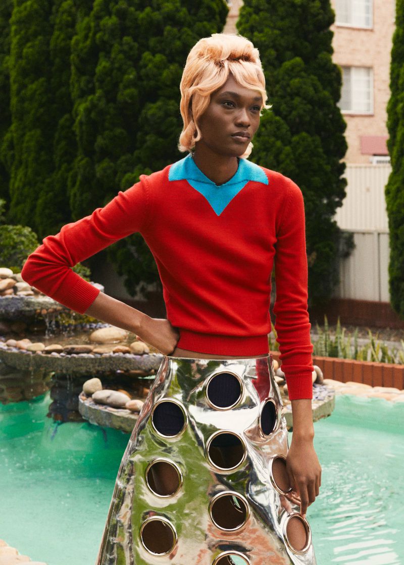 A woman with an orange hairdo in a red shirt and silver skirt is standing outdoors near a body of water in a fashion-style photograph.