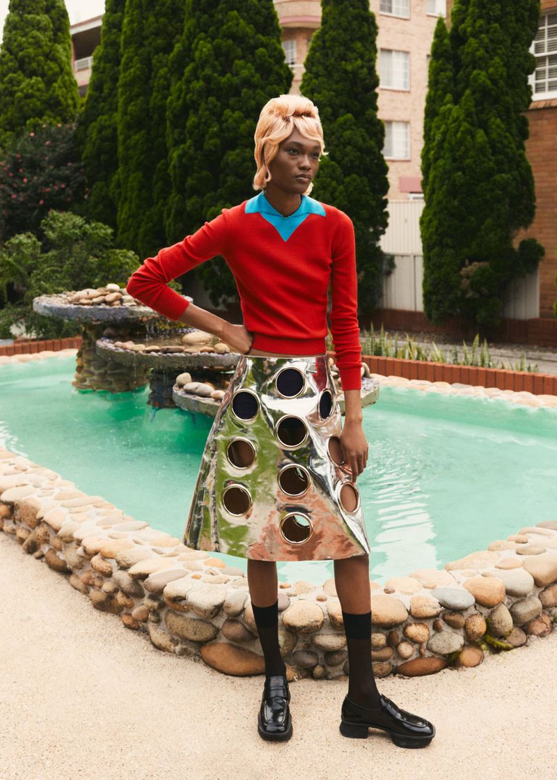 Outdoor fashion shot of a Black female model in a red sweater and silver skirt standing near a fountain in front of green trees.