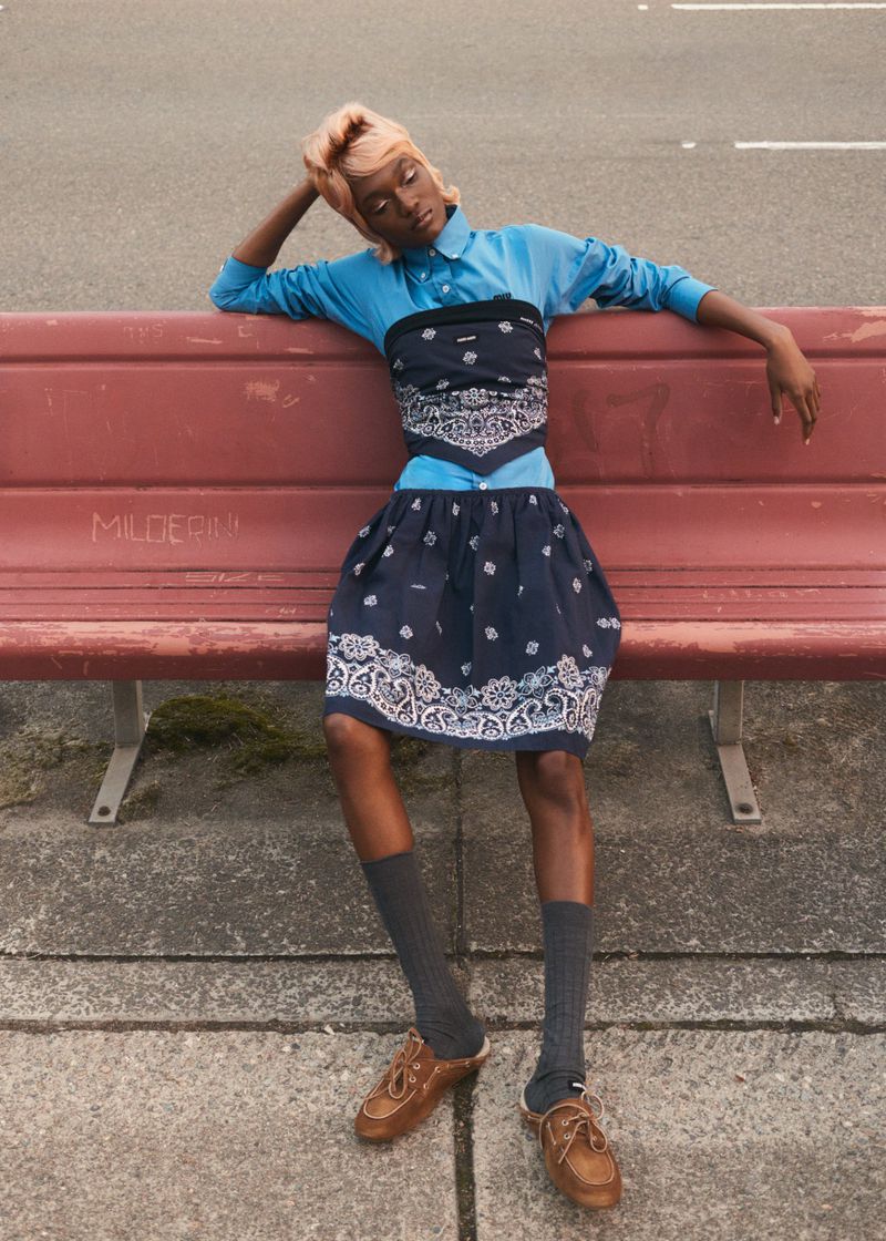 Woman sitting on bench wearing bandana-themed clothing. Young adult with tan skin, neutral expression, light brown shoes, and grey socks.