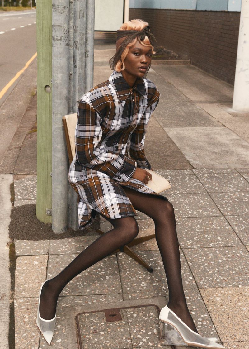 Stylish woman with blonde hair sits on a chair on a city street wearing a plaid dress, black tights, and translucent heels.