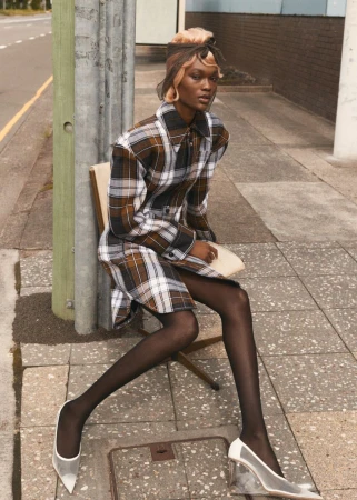 Stylish woman with blonde hair sits on a chair on a city street wearing a plaid dress, black tights, and translucent heels.
