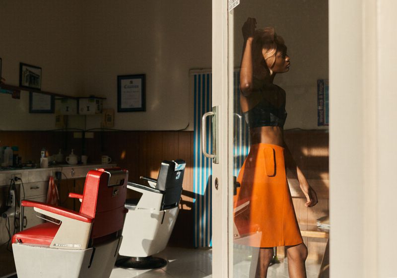 Woman in a barber shop with a black top and orange skirt, reflected in glass.