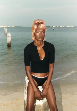 Woman with short blonde hair wearing black swimwear and top sitting on a stone block on a beach with ocean and horizon in the background.