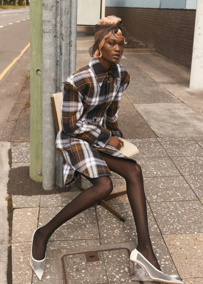 Model Naki Depass seated on an urban sidewalk wearing a brown plaid coat dress, black tights, and white wedge heels.