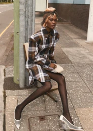 Model Naki Depass seated on an urban sidewalk wearing a brown plaid coat dress, black tights, and white wedge heels.