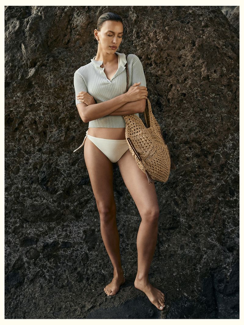 Model Irina Shayk wearing a ribbed polo shirt and bikini bottoms holding a woven straw tote against a rocky coast backdrop.