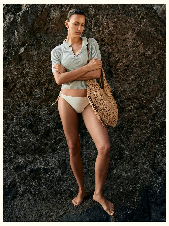 Model Irina Shayk wearing a ribbed polo shirt and bikini bottoms holding a woven straw tote against a rocky coast backdrop.