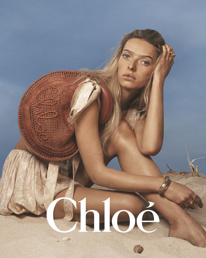 Model seated on sand against a clear sky holding a woven brown straw handbag in a Chloé campaign image