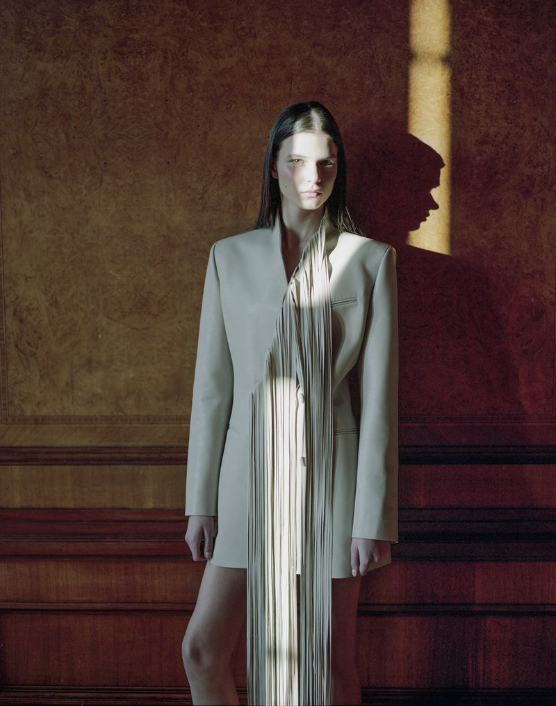 Portrait of a young woman with long dark hair in a beige jacket dress with a vertical fringe design against a paneled wall.