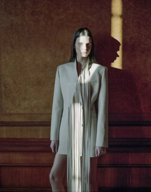 Portrait of a young woman with long dark hair in a beige jacket dress with a vertical fringe design against a paneled wall.
