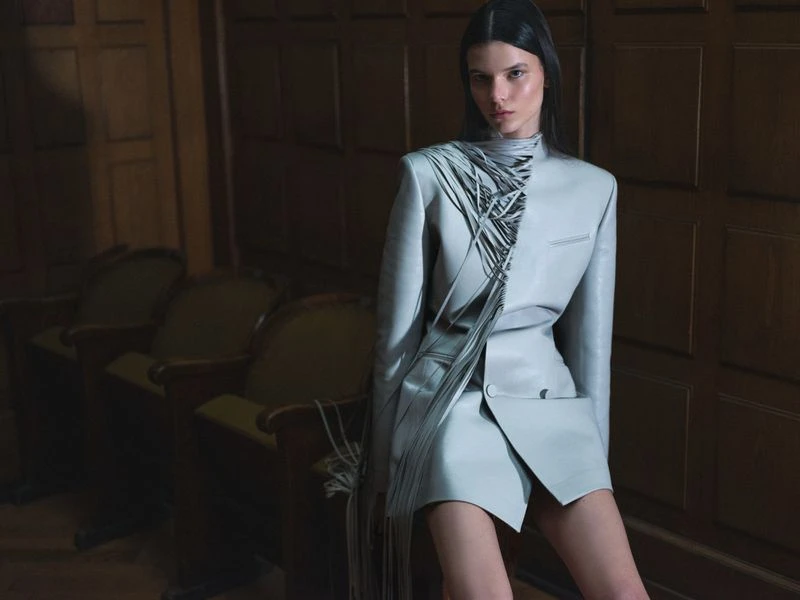 A brunette woman in a light blue jacket and skirt poses in a room with wood paneling and green upholstered chairs in soft lighting.