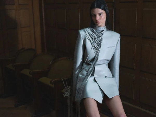 A brunette woman in a light blue jacket and skirt poses in a room with wood paneling and green upholstered chairs in soft lighting.