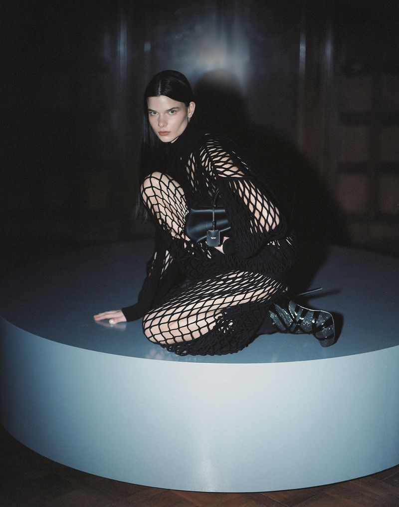 An adult woman in a black net-like dress squats on a light blue platform in a dimly lit room, showcasing a modern fashion aesthetic.