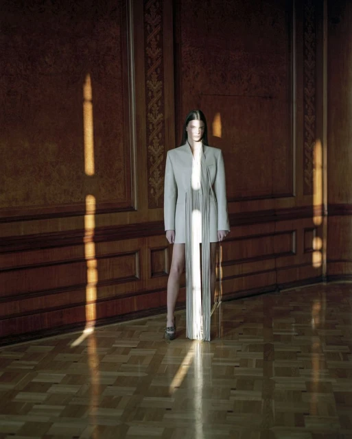 Elegant fashion shot: A slender female model in a light-colored blazer dress stands in a room with patterned wood floors and panelled walls.