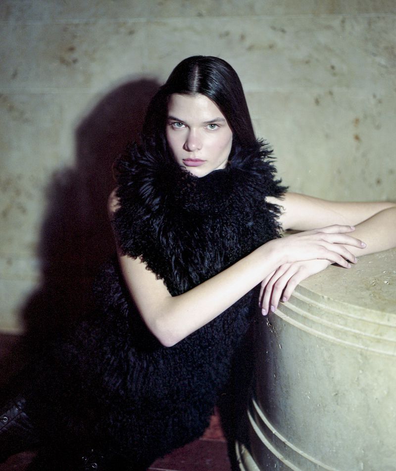 A woman with long dark hair poses in a black outfit with a large furry collar, leaning on a round stone structure indoors.