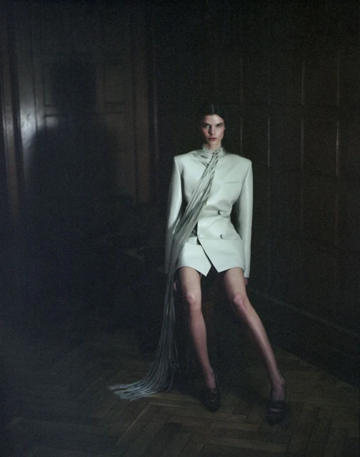 Fashion shot of woman in a pale-green skirt suit with a fringed detail posing while sitting indoors in front of a dark panelled wall.