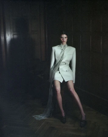 Fashion shot of woman in a pale-green skirt suit with a fringed detail posing while sitting indoors in front of a dark panelled wall.