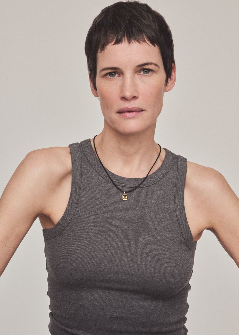 Close-up portrait of a woman with short dark hair wearing a grey ribbed tank top and a gold pendant necklace against a light grey backdrop.