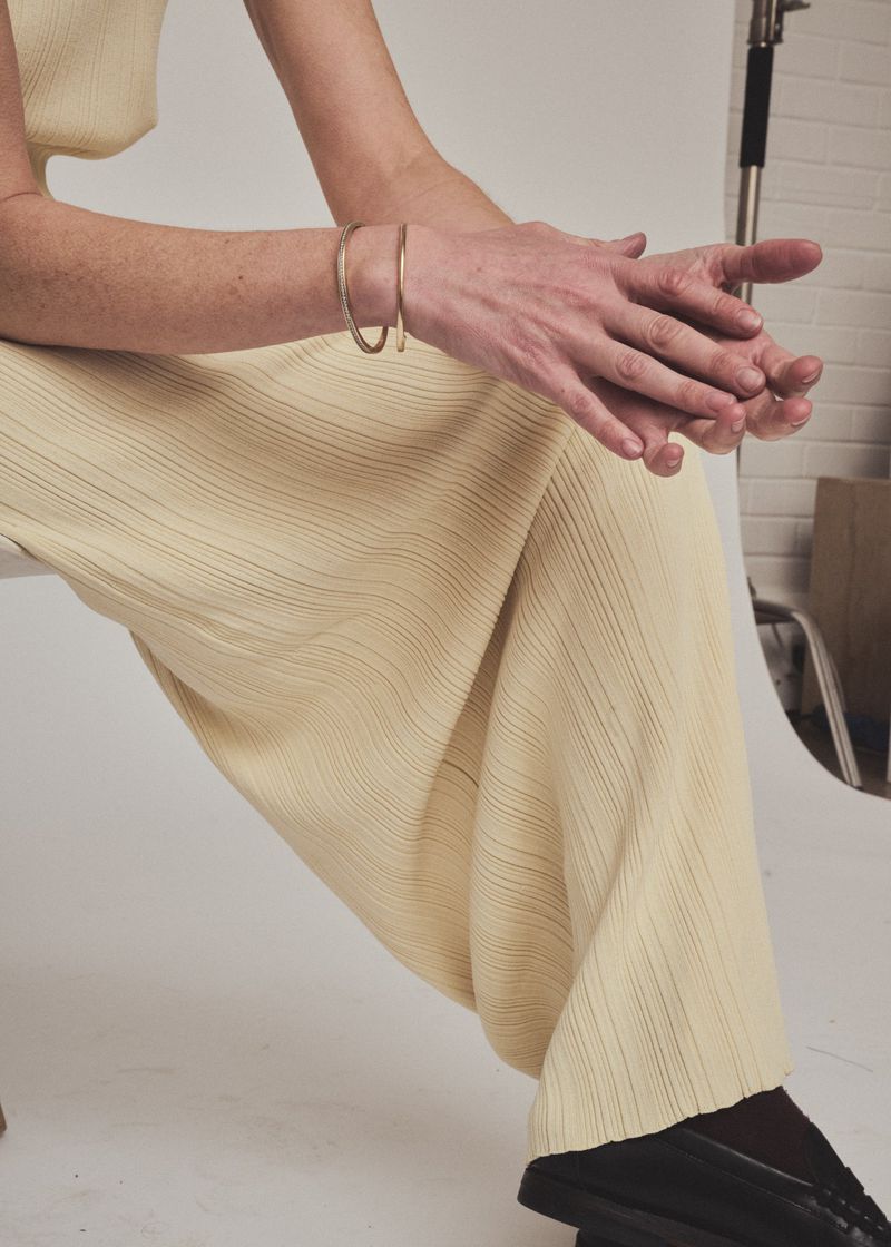 Close-up portrait of a seated woman wearing a ribbed yellow dress and gold bangles against a white studio seamless background.