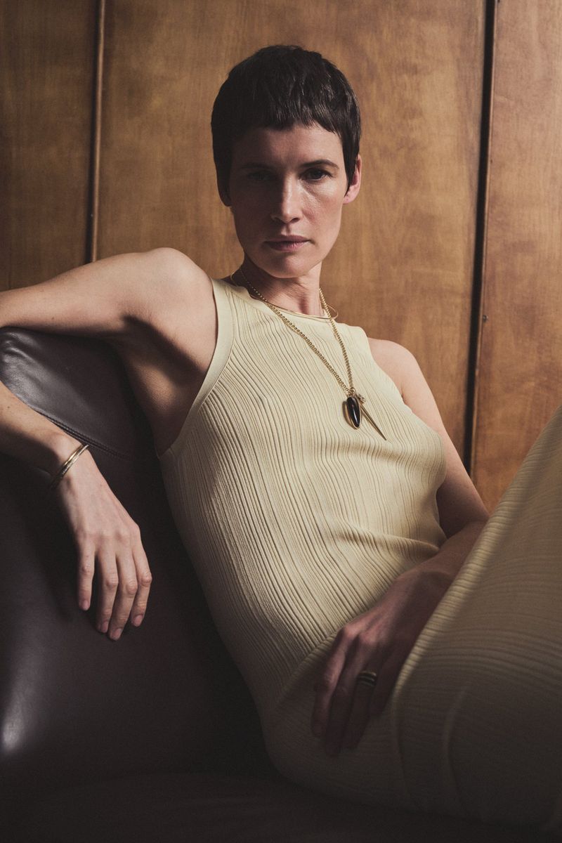 Short-haired woman in pale yellow ribbed dress wearing layered pendant necklaces seated on a leather chair against wooden paneling.
