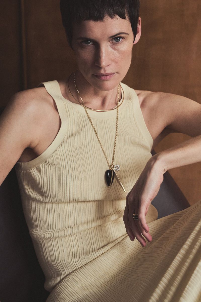 Woman wearing cream ribbed tank top and layered gold necklaces with pendant against textured warm brown background