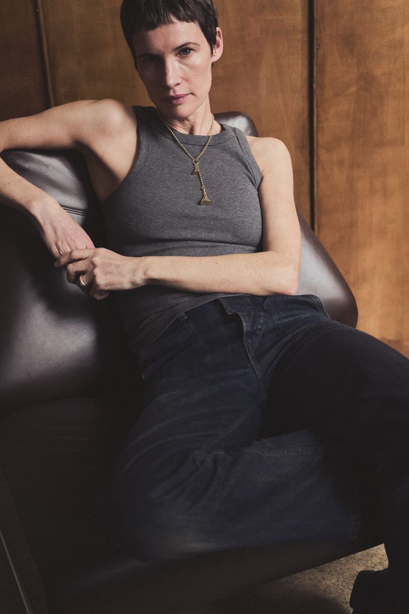Adult woman with short hair sits in a leather chair wearing a gray tank top and black pants in an indoor setting.