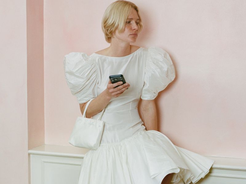 Blonde woman in a white puff-sleeve bridal dress holding a small white handbag and a smartphone against a pale pink wall.