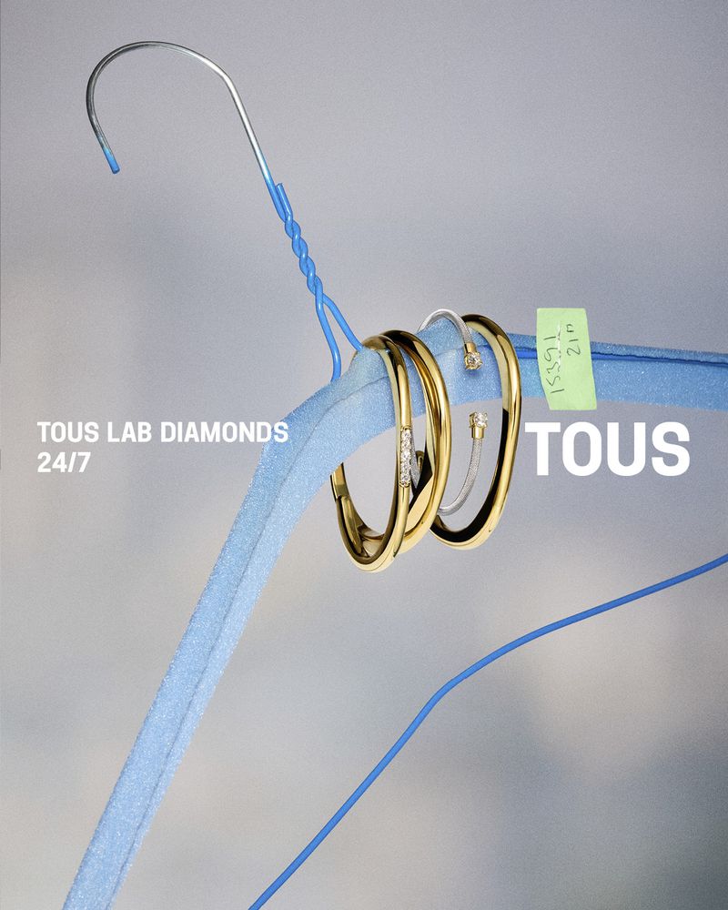 Gold bangles and rings hanging on a blue padded coat hanger against a gradient grey background for Tous Lab Diamonds