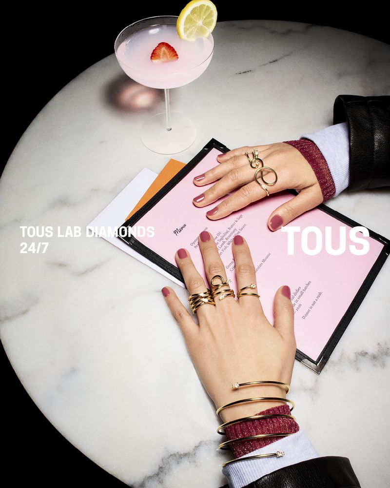 Close-up of hands adorned with gold rings and bracelets resting on a pink menu next to a cocktail on a round marble table