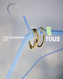 Gold bangles and rings hanging on a blue padded coat hanger against a gradient grey background for Tous Lab Diamonds